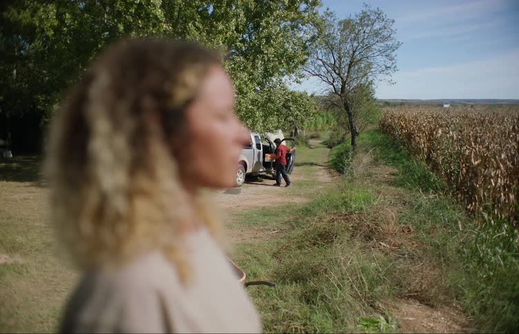 Woman stands looking out toward cornfield as man arrives with wrapped gifts