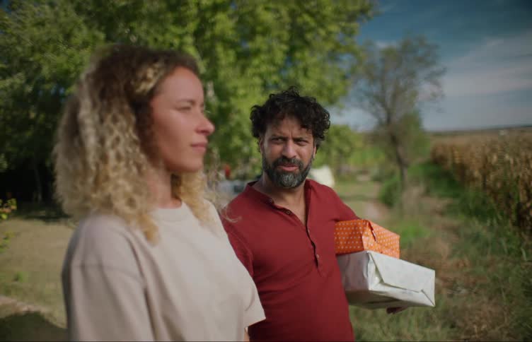 A man approaches a woman outside with wrapped gifts under his arm