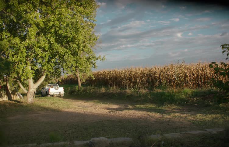 A woman sits in the bed of a truck as it pulls off down a dirt road in a rural area