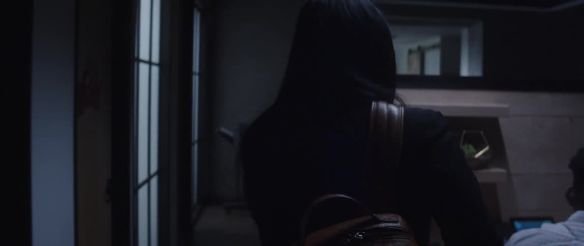 A woman walks through a door and into a room where a man is sitting in front of a computer and she speaks to him