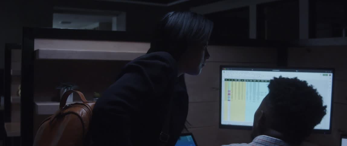 A man is sitting at a desk with computers as a woman stands next to him looking at the monitor