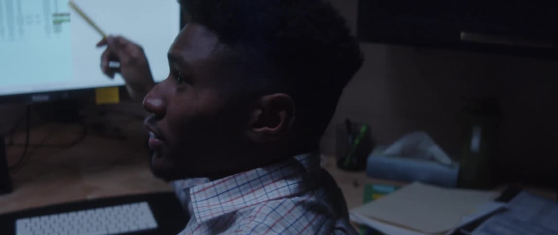 A man is sitting at a desk with a laptop computer and a monitor as he holds a pen in his hand pointing at the monitor as he speaks to a woman standing next to him