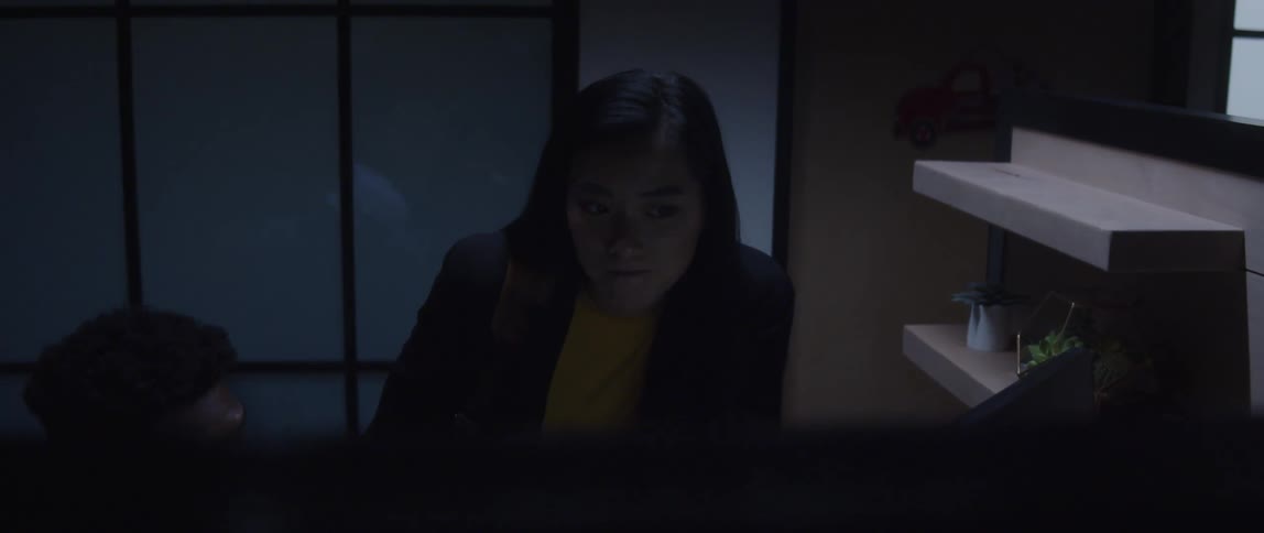A man is sitting at a desk with a computer as a woman stands next to him looking at the monitor as she talks to him