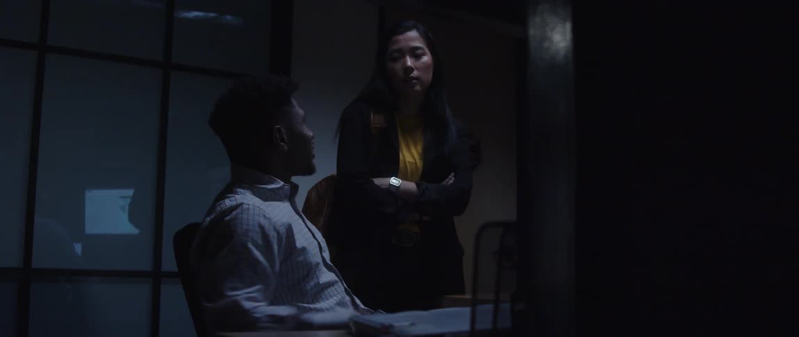 A man is sitting at a desk with a computer as he talks to a woman standing next to him