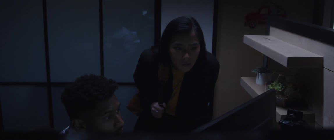 A man is sitting at a desk in front of a computer as a woman stands near him looking at the monitor as he works