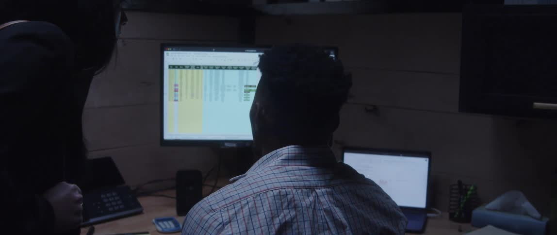 A man is sitting at a desk in front of computers as a woman stands next to him looking at the monitor as they talk and point at the screen