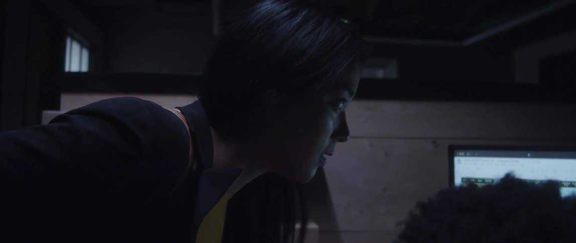 A man is sitting at a desk with computers as a woman stands next to him looking at the monitor as she talks to him