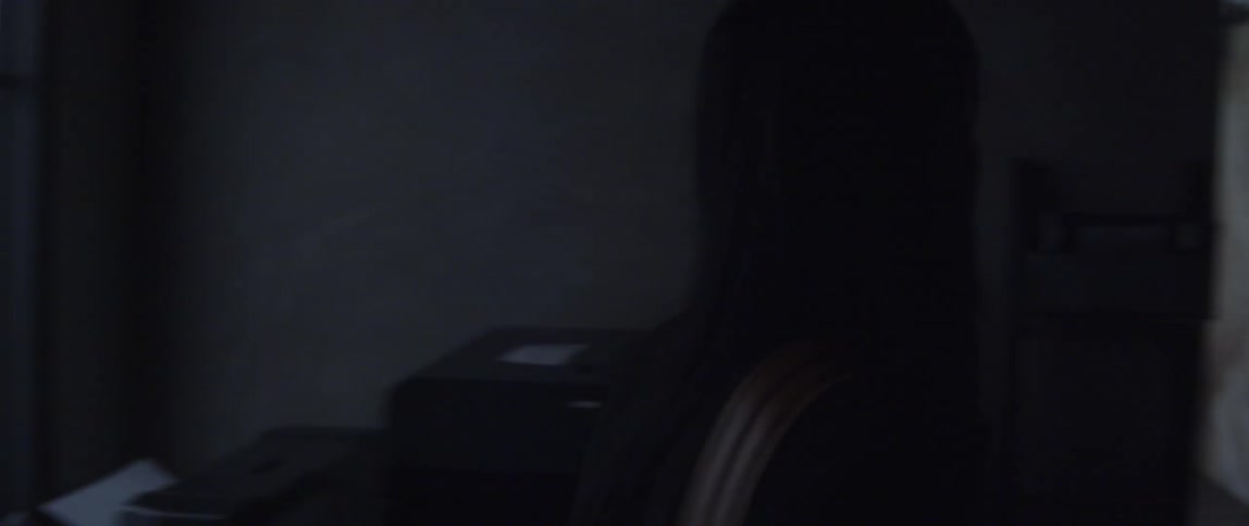 A woman is standing in a dark room as she says goodbye and waves as she walks out through the hallway towards the exit