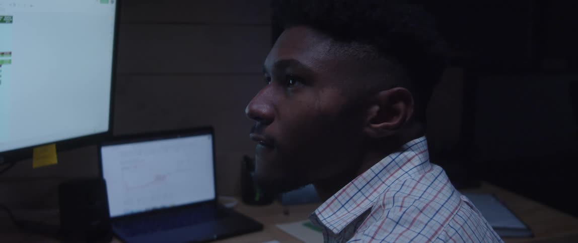 A man is sitting at a desk with a laptop computer and a large monitor with his head turned as he is thinking