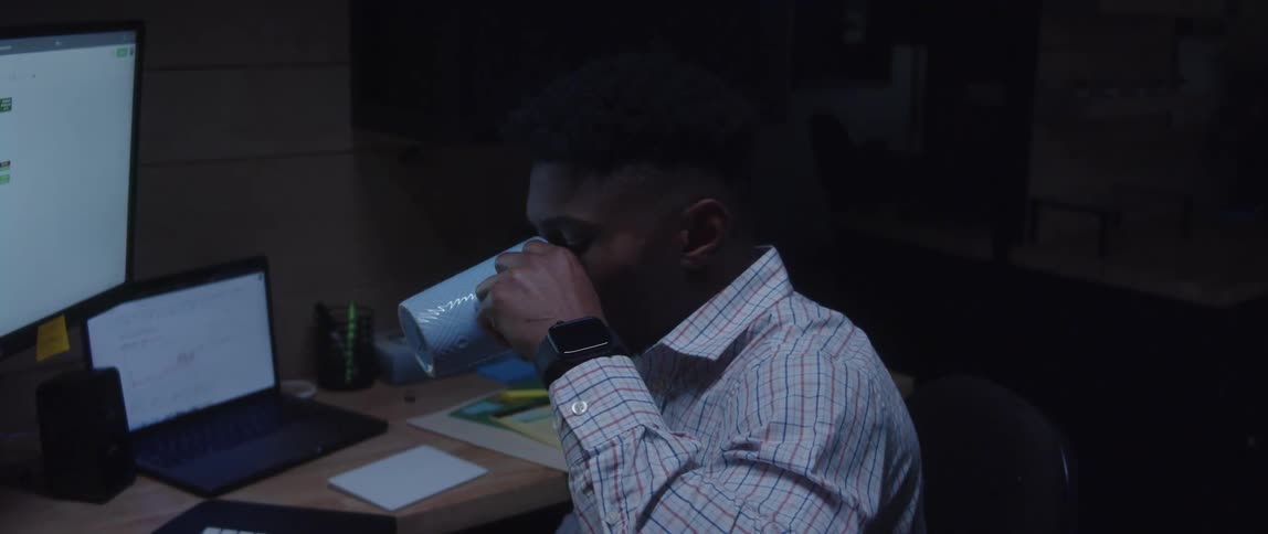 A man sitting at his desk drinking out of a blue mug and looking at his computer