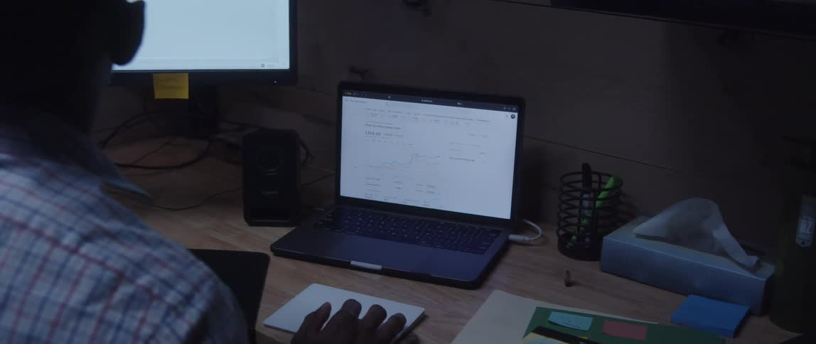 A young black man sits at an office desk while comparing information between a laptop screen and a larger computer monitor