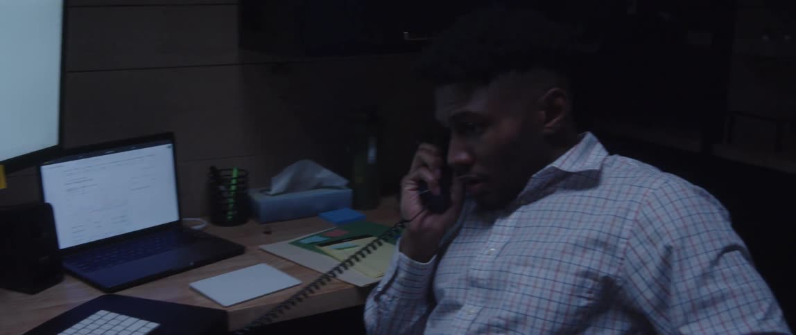 A man is sitting at a desk in front of computers as he talks on the telephone