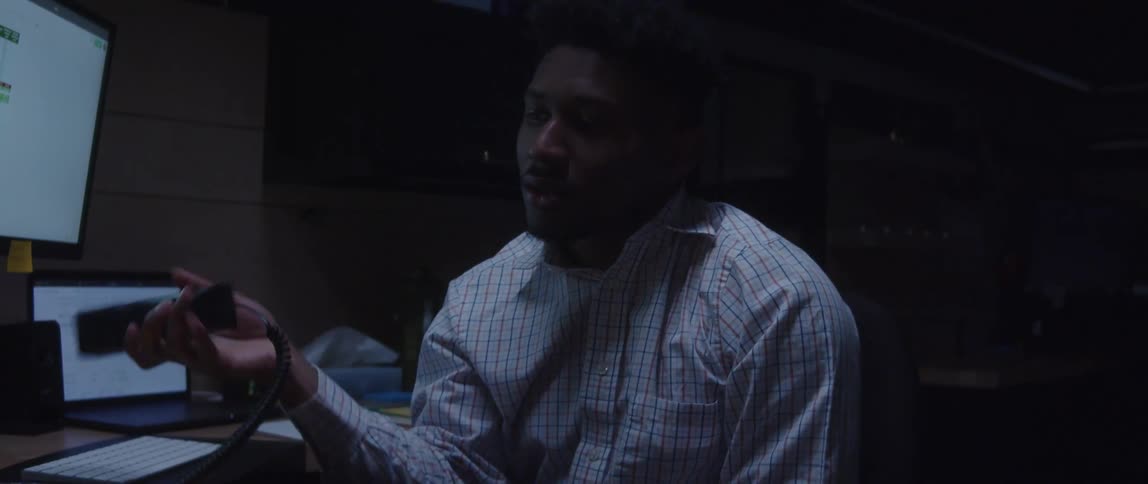 A man is sitting at a desk with computers as he talks on the phone and then hangs up the receiver in frustration