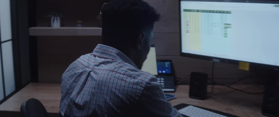 A man is sitting at a desk looking at a large monitor with a laptop computer next to him as he picks up a notepad