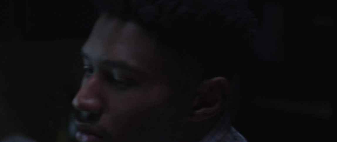 A black man sits at a work desk with a computer monitor and answers the phone