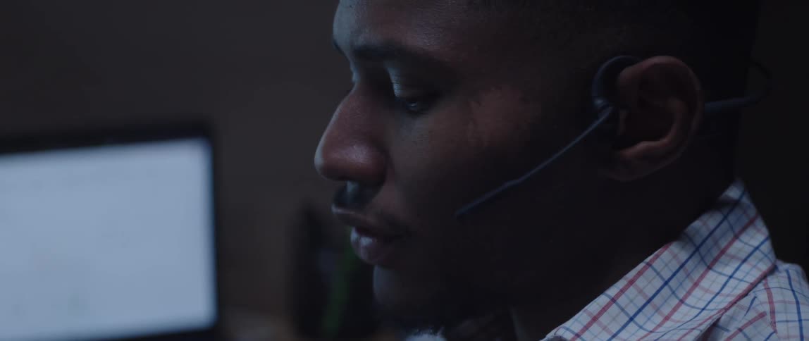 A man in an office sits by a computer and talks on a headset