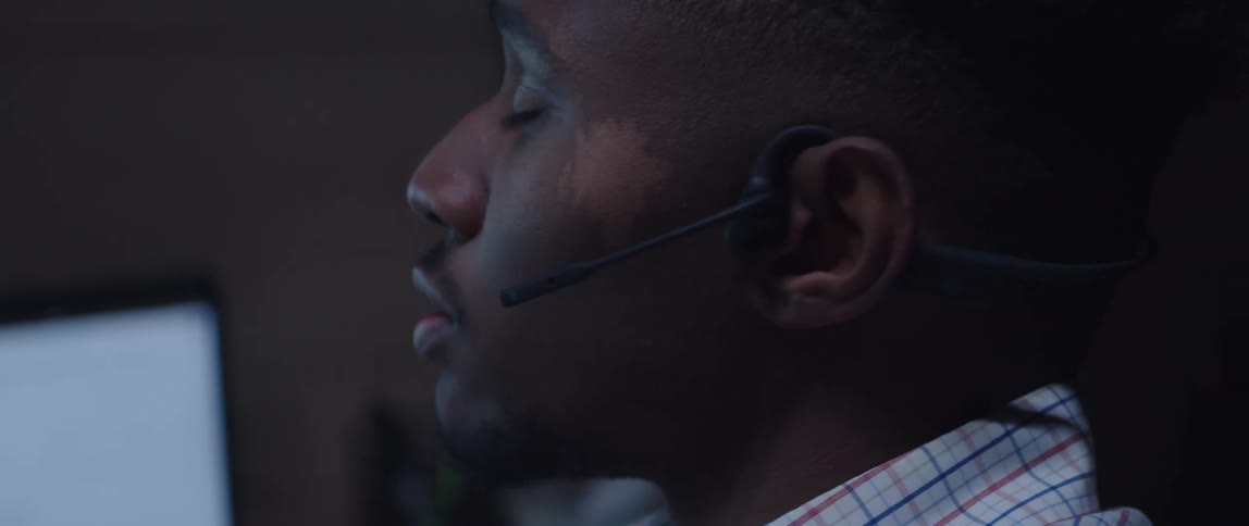 An up close of a mans face while he talks on an ear piece and then points to his computer screen
