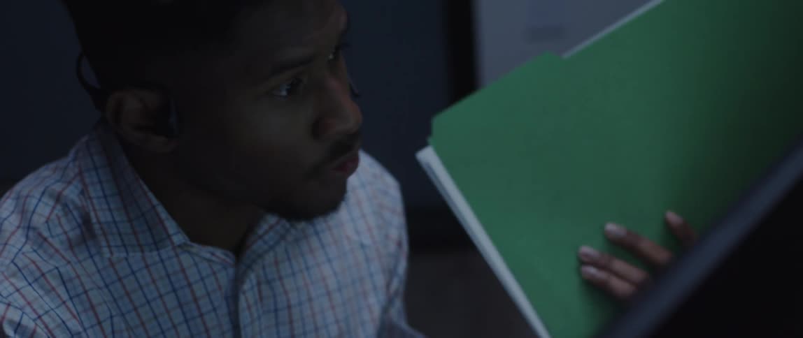 A man is talking on the phone sitting at a desk holding a folder in his hand and looking up as he pauses to think