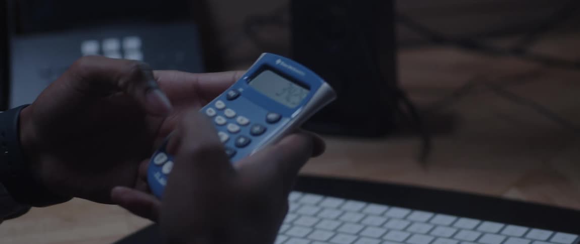 A man is sitting at a desk looking at a chart on a computer monitor and using a small portable calculator