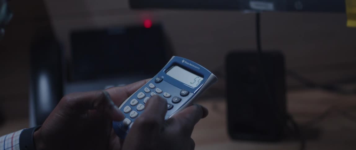 A man is sitting at a desk using a calculator as he points to a chart on the computer monitor