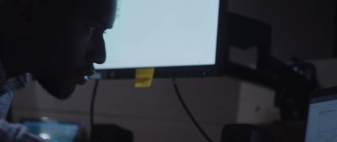 A man is sitting at a desk looking at a monitor and then looking over at a laptop computer as he works