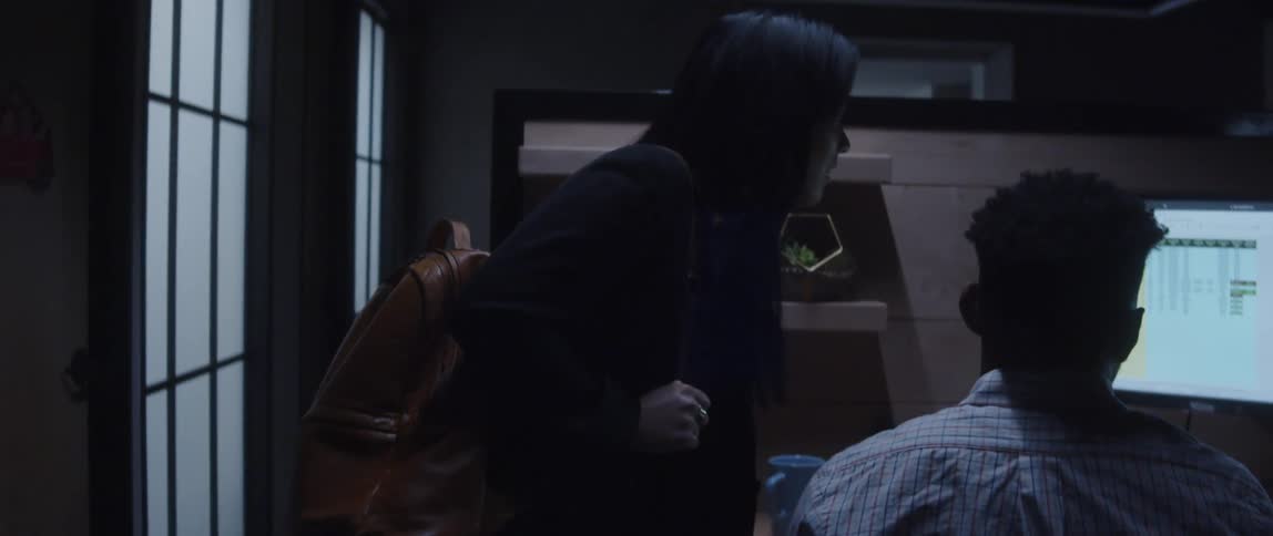 A man sitting in front of his computer desk while a woman with a backpack on leans over and observes his screen as they converse