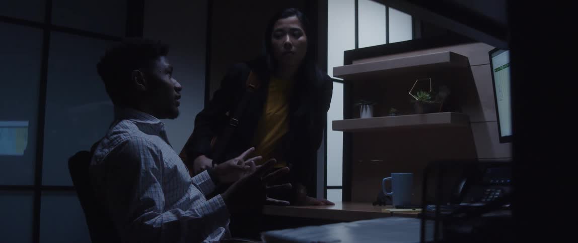 A man sitting at his desk talking to a women leaning on his desk nodding her head at him