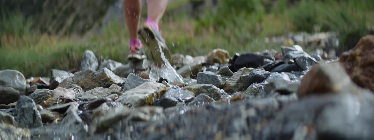 A stream flows through rocks and a girl splashes water as she runs through to the grass