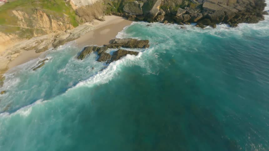 Rocks are scattered along the beach near a cliff as waves roll into shore and the reflection of the sun sparkles on the water