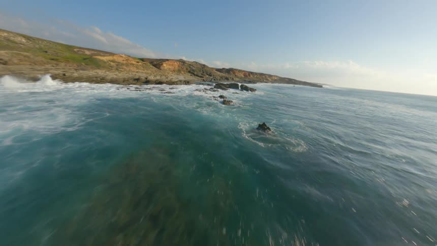 The sun shines brightly upon the water as waves roll into shore and splash against rocks along the coast