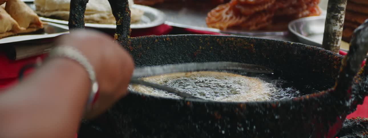 Woman uses metal tongs to move dough frying in metal pan on grill