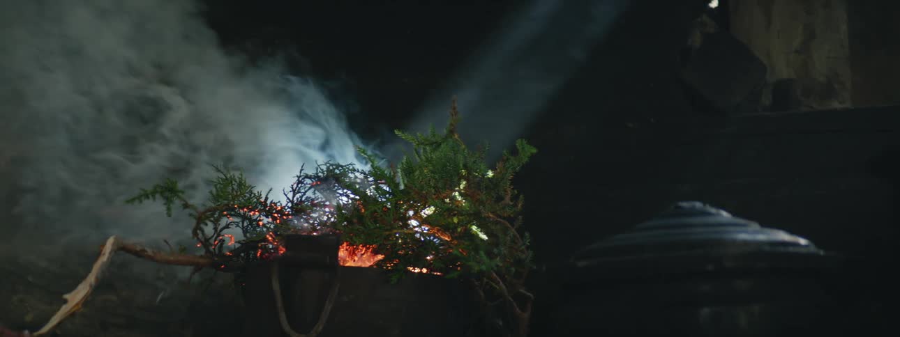 A branch burning over a fire in a room with a pot near it and smoke rolling off the fire
