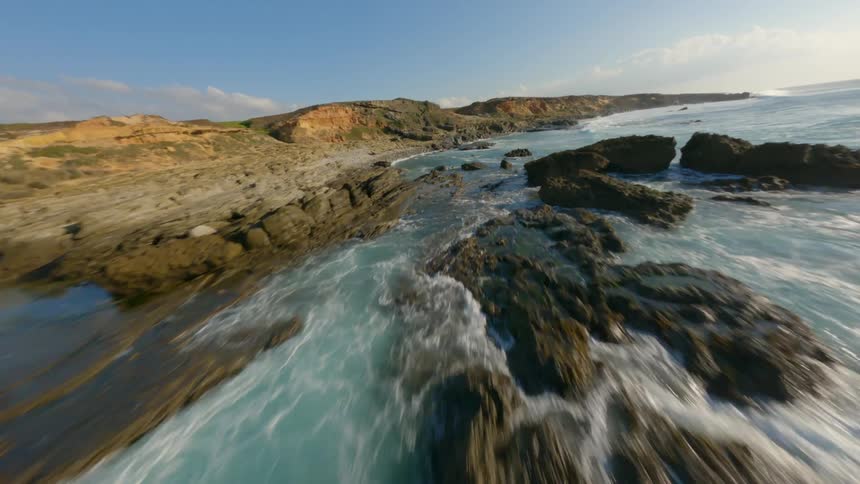 Moving across the coast as blue water sparkles in the sunlight as waves roll in to shore and splash against rocks and roll over the shore