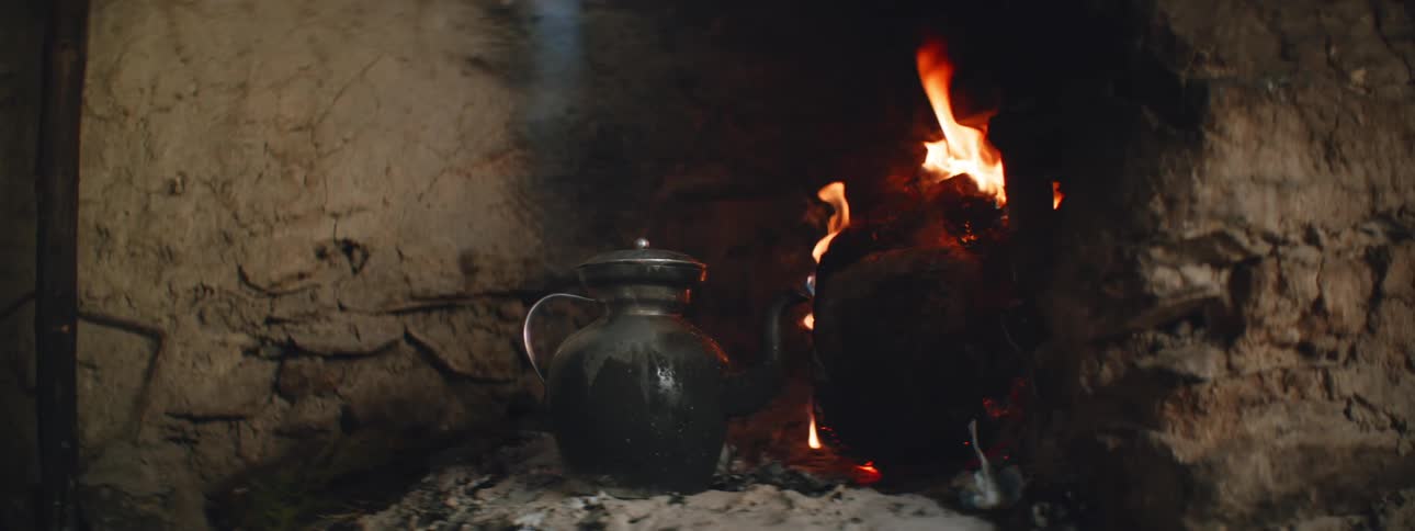 The flames of a fire burn brightly inside a cave as a pot sits on rocks near the fire