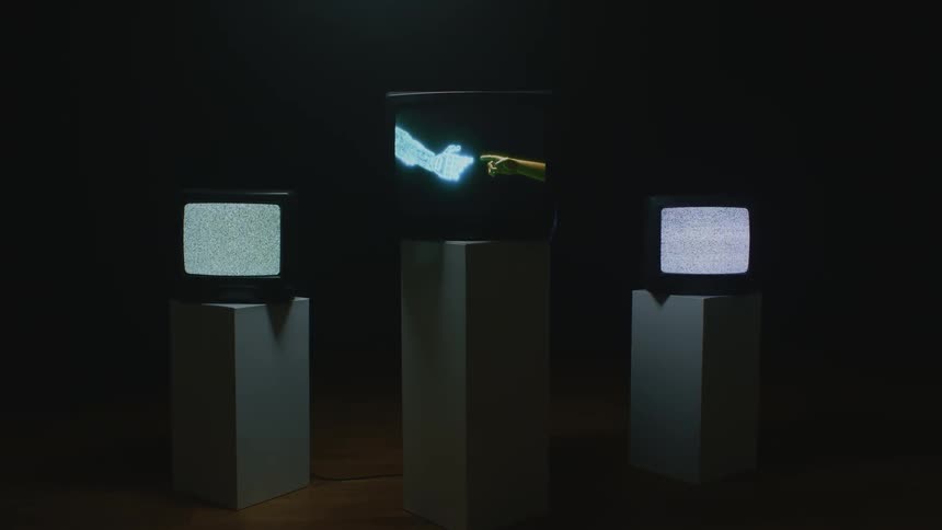 Three televisions on pedestals two with a static screen and one with two hands reaching eachother