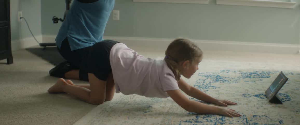 A woman and young girl lean over on the floor and stretch their arms straight out then up high as they watch a tablet video