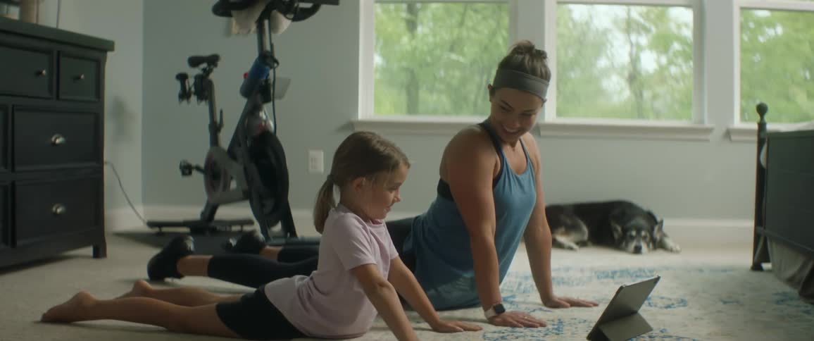 Mother and daughter exercise on bedroom rug using tablet while pet dog watches