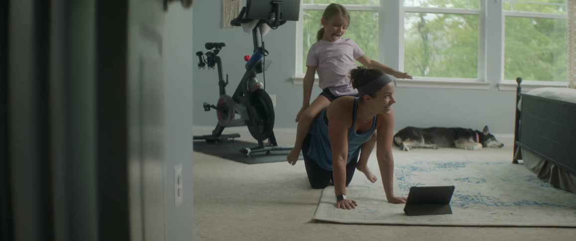 Woman on her hands and knees as a little girl sits on womans back dog sleeping in back ground under window and next to exercise bike