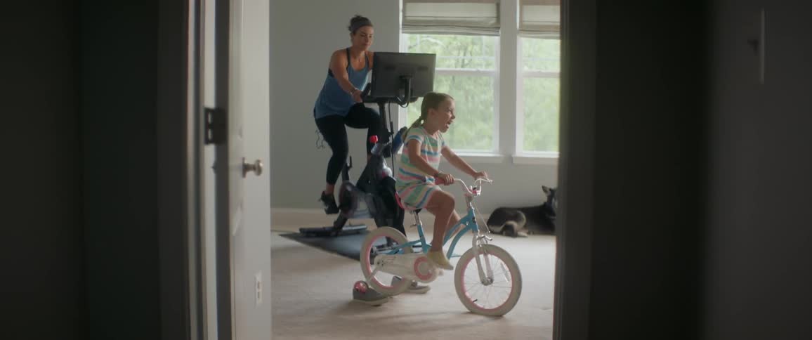 A woman on an exercise bike smiles at a girl peddling a childrens bike with its back wheel suspended in the air beside her