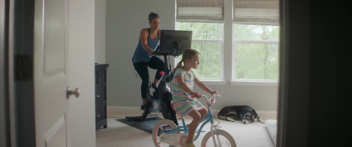 A mom and daughter exercise on stationary bikes indoors with a dog on the floor next to them