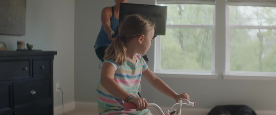 A young girl pedels a stationary bike inside with her mother in the background riding an exercise bike