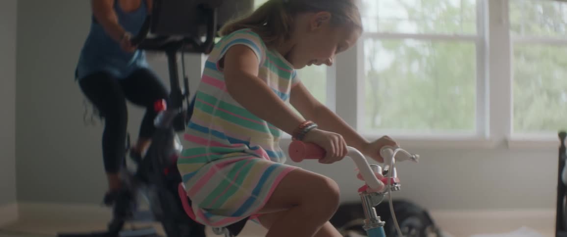 A girl and mom ride stationary bikes indoors during the day with a dog on the floor in the same room
