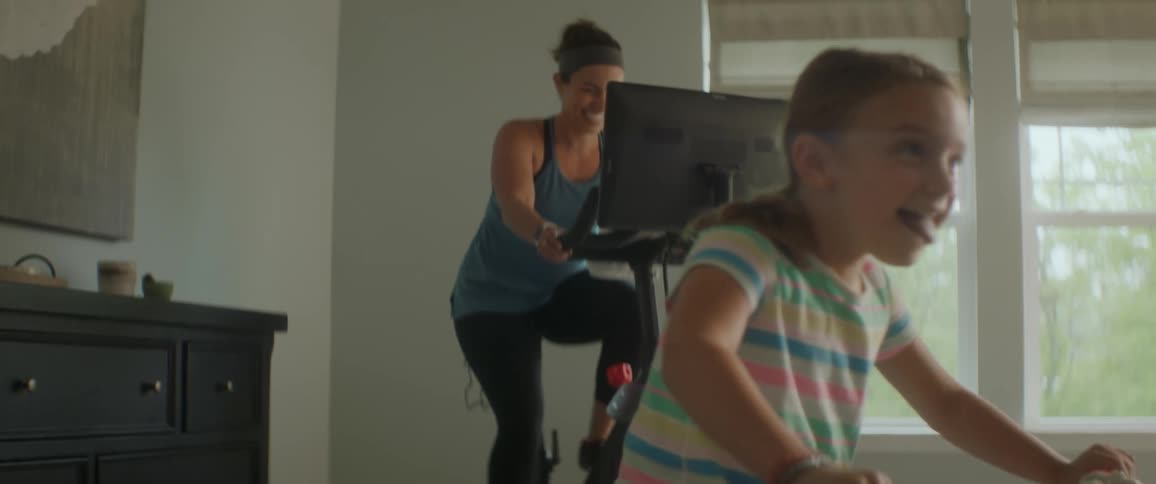 A woman rides a computerized exercise bike with her daughter riding a bicycle in the same room