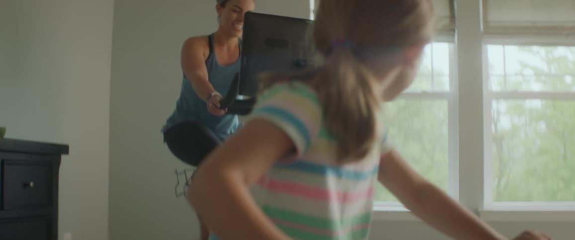 A woman riding a smart exercise bike in a living room smiles at a young girl riding a childrens exercise bike in front of her