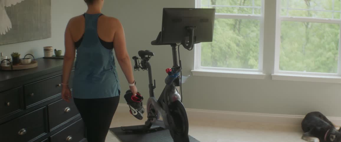 A woman walks over to a stationary bike carrying sneakers in a room during the day