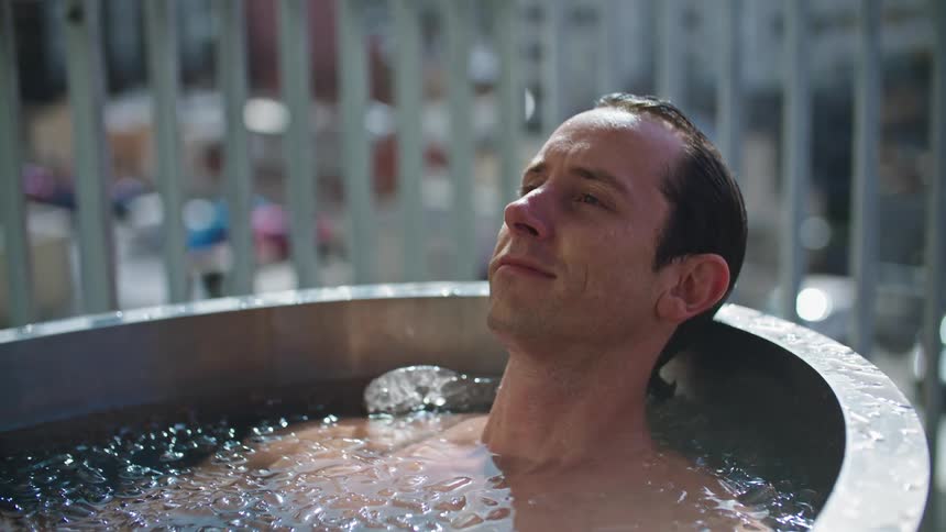 A man is leaning back as he sits in outside in a bathtub and he smiles