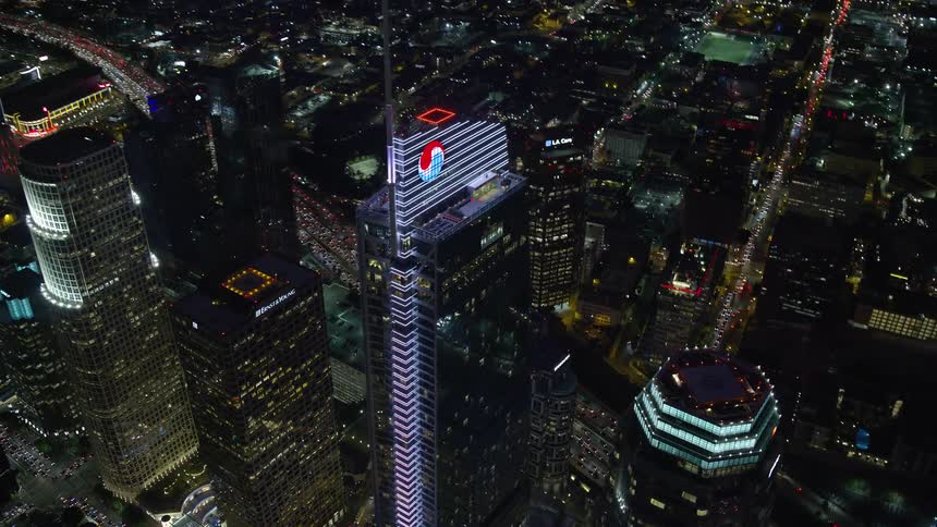 An aerial view of Wilshire Grand Center a modern glass building shot at night from an approaching orbiting and tilting -- surrounded by other downtown LA skyscrapers -- in superstructures-filled Southern California during spring or summer as seen from a s