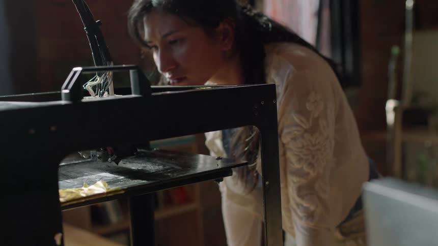 Woman leans forward watching 3d printer near windows in brick walled room
