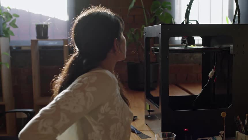 A woman sitting at a desk holding an object in her left hand and looking at the machine next to her