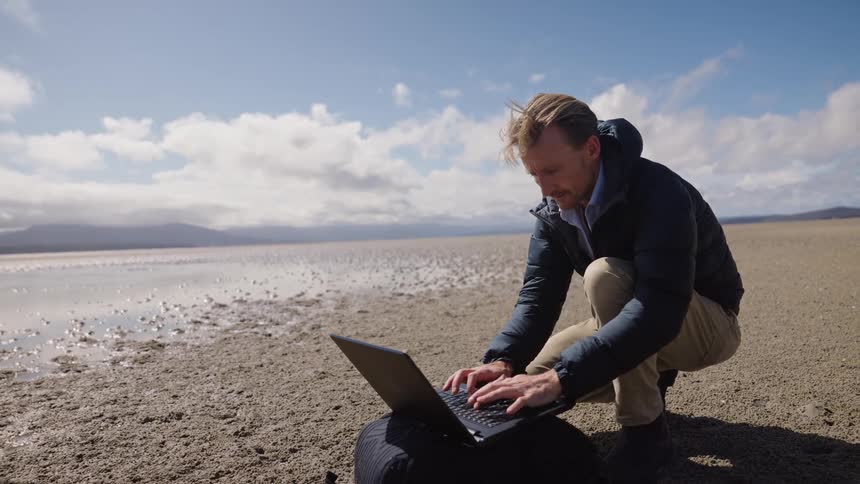 A man kneeling in front of his laptop that sits on a bag on the sandy ground as the man looks around and then types on the laptop